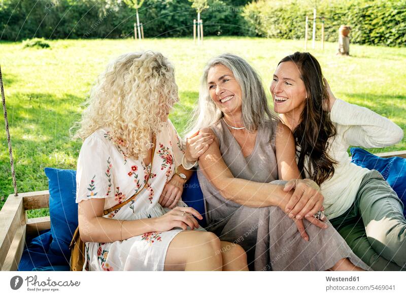 Drei glückliche Frauen unterschiedlichen Alters sitzen auf einem Hängebett im Garten und unterhalten sich sprechen reden entspannt entspanntheit relaxt sitzend