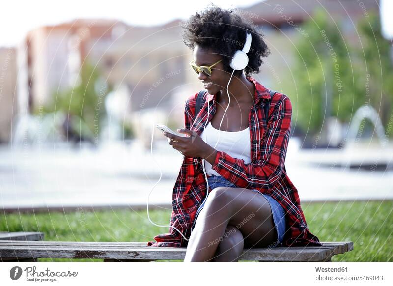 Lächelnde junge Frau sitzt auf einer Bank im Stadtpark und hört Musik mit Kopfhörern Smartphone iPhone Smartphones hören hoeren sitzen sitzend lächeln weiblich