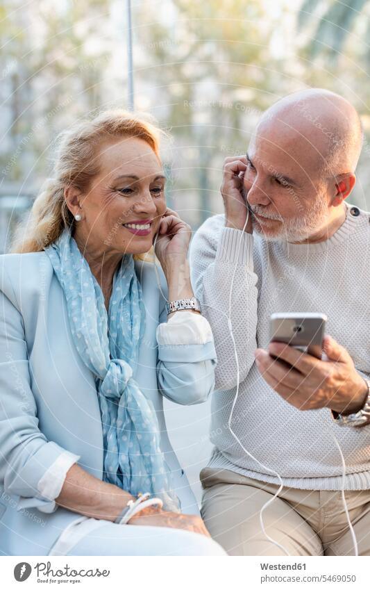 Spanien, Barcelona, glückliches älteres Paar, das sich ein Smartphone mit Ohrstöpseln teilt Ohrhörer Teilen Sharing Pärchen Paare Partnerschaft Glück