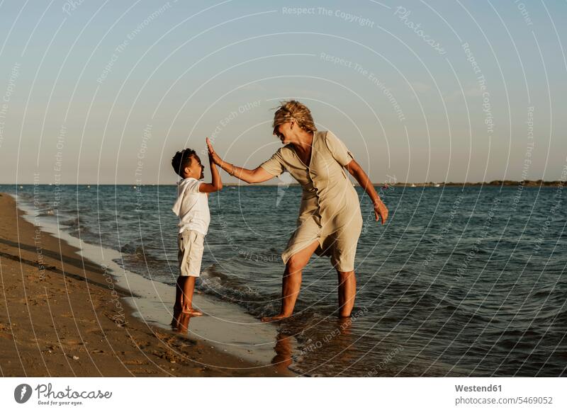 Glückliche Großmutter und Enkel, die sich bei Sonnenuntergang am Strand gegenseitig das High Five geben Farbaufnahme Farbe Farbfoto Farbphoto Außenaufnahme
