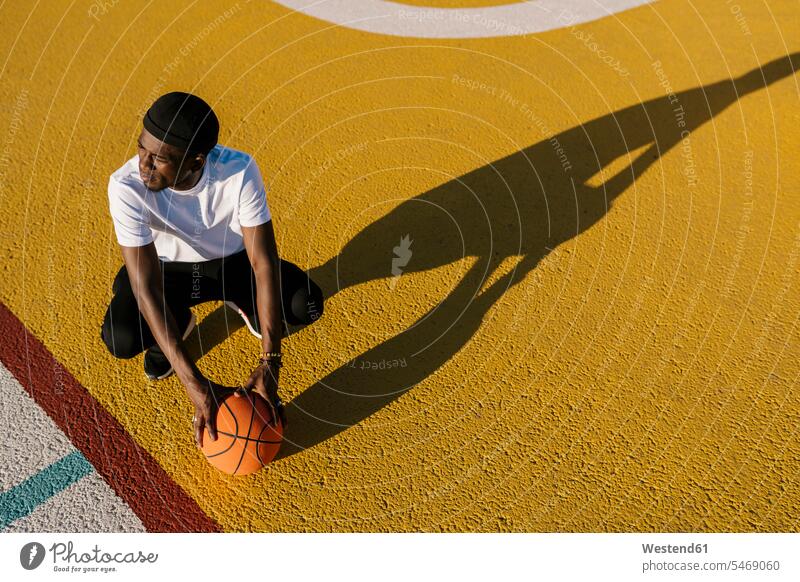 Nachdenklicher junger Mann hält Basketball an einem sonnigen Tag auf dem Sportplatz kauernd Farbaufnahme Farbe Farbfoto Farbphoto Spanien Freizeitbeschäftigung