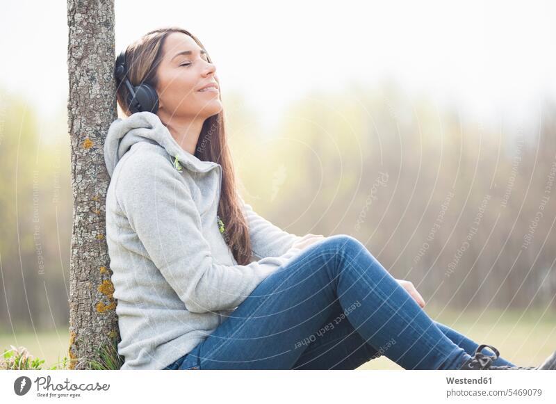 Entspannte Frau hört Musik, während sie sich im Park an einen Baumstamm lehnt Farbaufnahme Farbe Farbfoto Farbphoto Außenaufnahme außen draußen im Freien Tag