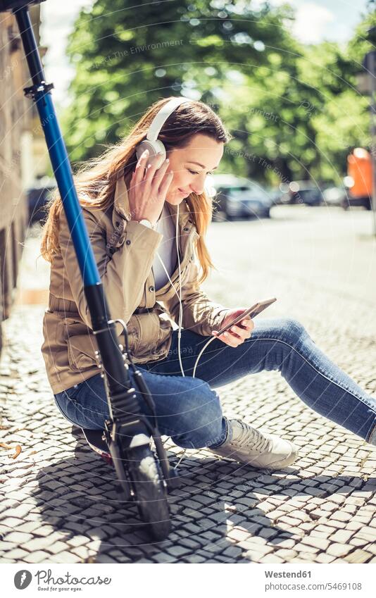 Lächelnde Frau sitzt auf einem E-Scooter und hört Musik mit Kopfhörern und Smartphone Kopfhoerer Telekommunikation Handies Handys Mobiltelefon Mobiltelefone