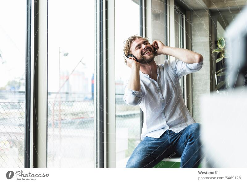 Glücklicher lässiger junger Mann, der mit Kopfhörern am Fenster Musik hört glücklich glücklich sein glücklichsein hören hoeren Kopfhoerer Männer männlich leger
