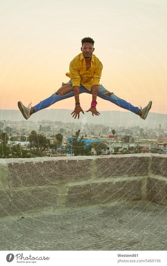 Junger Mann im gelben Hemd springt bei Sonnenuntergang von einer Mauer Hemden springen Spruenge Sprünge hüpfen abends begeistert Enthusiasmus enthusiastisch