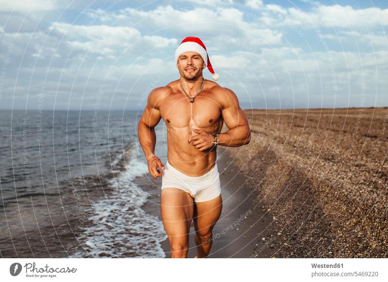 Lächelnder muskulöser junger Mann mit Nikolausmütze rennt am Strand gegen bewölkten Himmel Farbaufnahme Farbe Farbfoto Farbphoto Spanien Freizeitbeschäftigung
