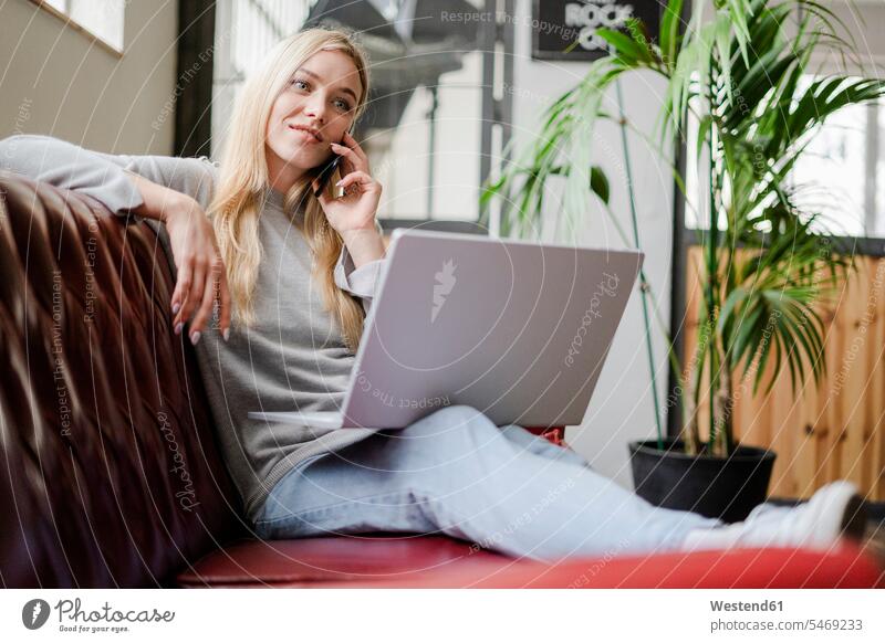 Lächelnde junge Frau sitzt auf Couch mit Laptop sprechen auf Handy weiblich Frauen telefonieren anrufen Anruf telephonieren sitzen sitzend lächeln Mobiltelefon