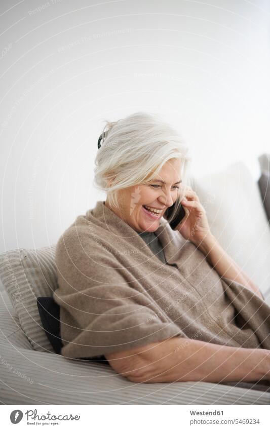 Fröhliche ältere Frau, die am Handy spricht, während sie gegen eine weiße Wand zu Hause sitzt Farbaufnahme Farbe Farbfoto Farbphoto Dänemark Skandinavien