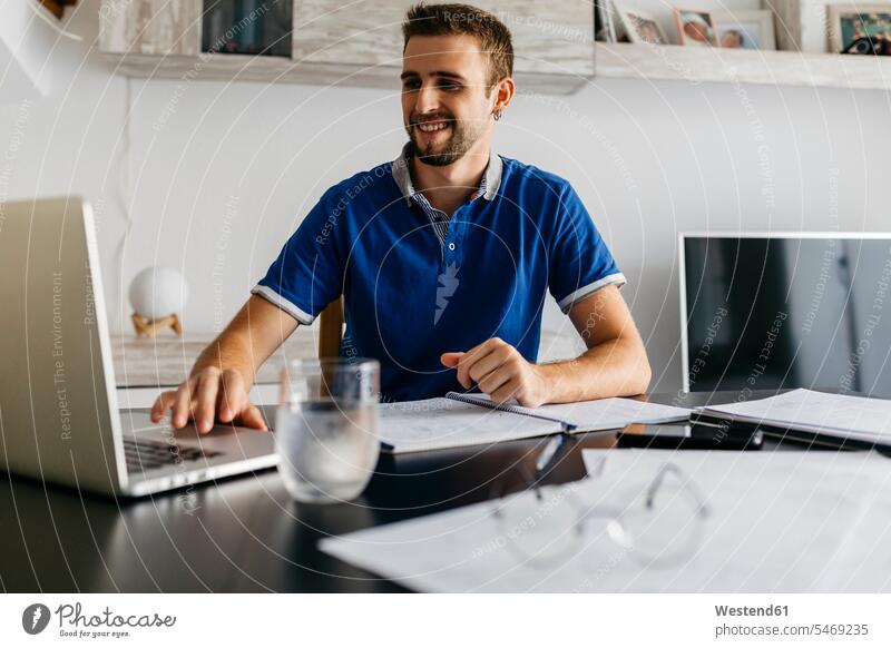 Lächelnder gutaussehender junger Mann mit Laptop bei den Hausaufgaben am Tisch Farbaufnahme Farbe Farbfoto Farbphoto Innenaufnahme Innenaufnahmen innen drinnen