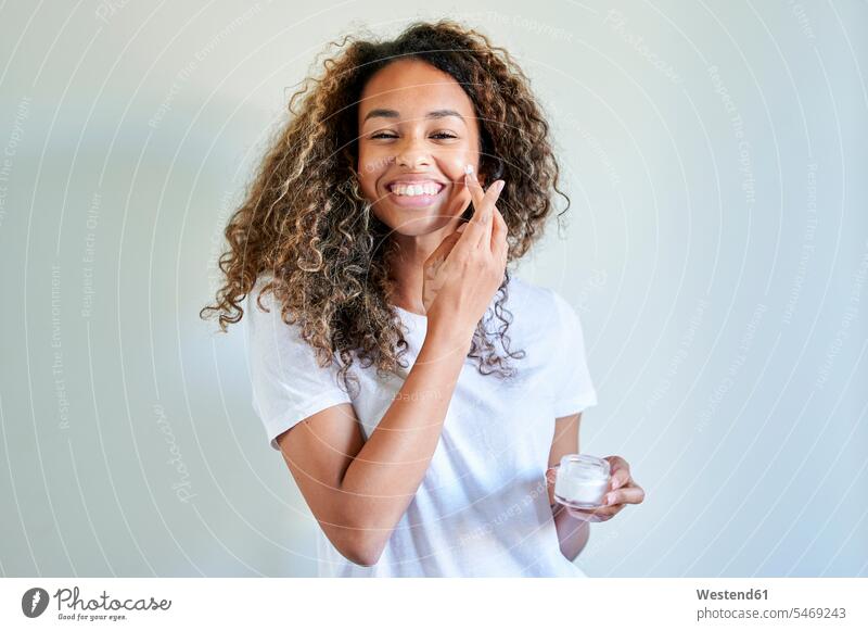 Fröhliche junge Frau, die Gesichtscreme gegen eine weiße Wand aufträgt Farbaufnahme Farbe Farbfoto Farbphoto Innenaufnahme Innenaufnahmen innen drinnen Spanien