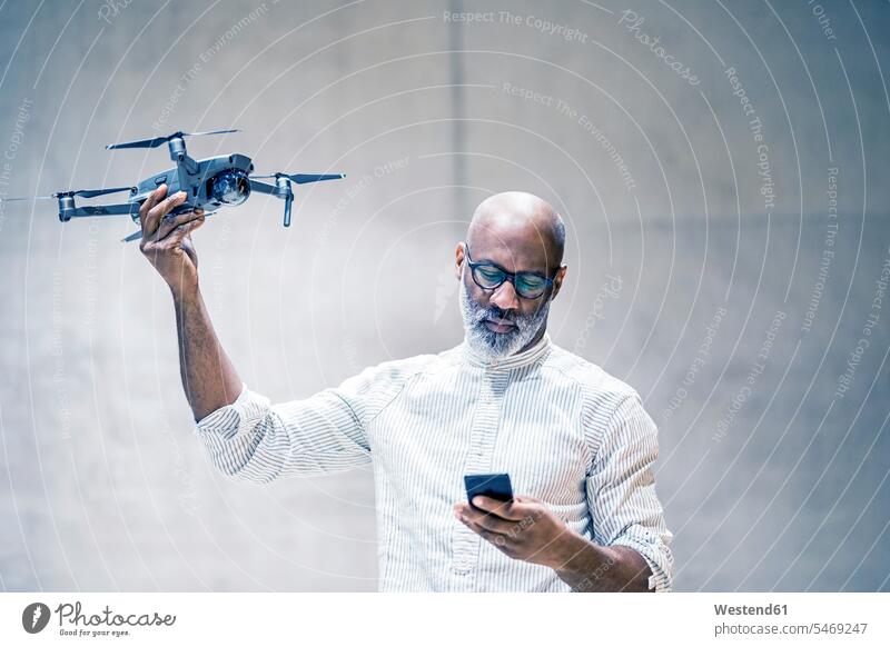 Porträt eines glatzköpfigen reifen Mannes, der einen Quadcopter hält, während er auf ein Mobiltelefon schaut Geschäftsmann Businessmann Businessmänner