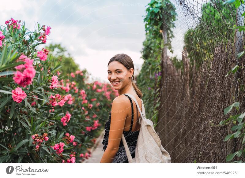 Porträt einer lächelnden jungen Frau mit Rucksack beim Spaziergang in einem Park Leute Menschen People Person Personen Europäisch Kaukasier kaukasisch