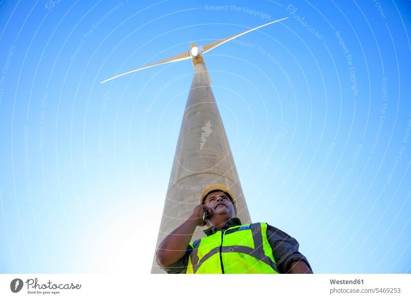Niedriger Blickwinkel des Technikers am Handy vor der Windkraftanlage Windturbine Windrad Windräder telefonieren anrufen Anruf telephonieren Mobiltelefon