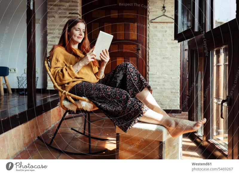 Lächelnde Frau benutzt digitales Tablet, während sie zu Hause auf einem Stuhl sitzt Farbaufnahme Farbe Farbfoto Farbphoto Innenaufnahme Innenaufnahmen innen
