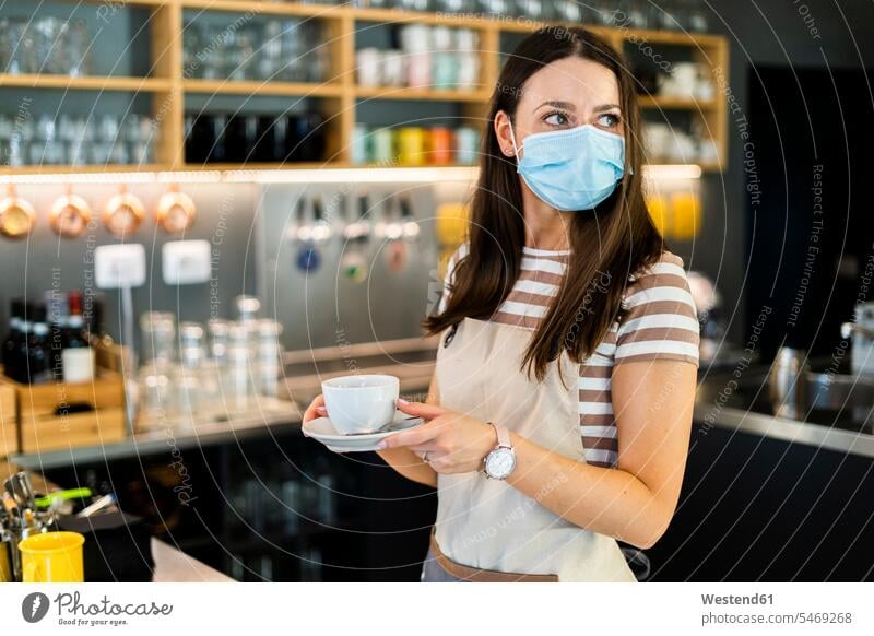 Nachdenklicher junger Besitzer trägt Maske, während er Kaffeetasse und Untertasse im Café hält Farbaufnahme Farbe Farbfoto Farbphoto Innenaufnahme