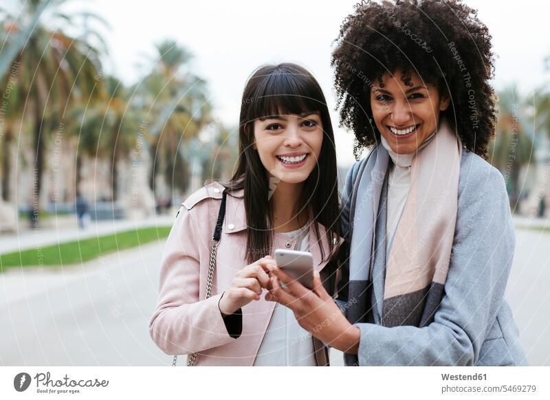 Spanien, Barcelona, Porträt von zwei glücklichen Frauen mit Handy auf der Promenade Portrait Porträts Portraits Promenaden Freundinnen Mobiltelefon Handies