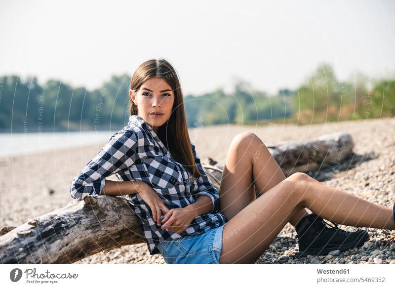 Junge Frau sitzt am Kieselsteinufer am Baumstamm sitzen sitzend weiblich Frauen Ufer Kieselsteine Holzstamm Holzstämme Erwachsener erwachsen Mensch Menschen