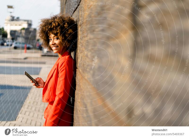 Porträt einer lächelnden jungen Frau mit Handy in modischem roten Hosenanzug an der Wand lehnend Telekommunikation Handies Handys Mobiltelefon Mobiltelefone