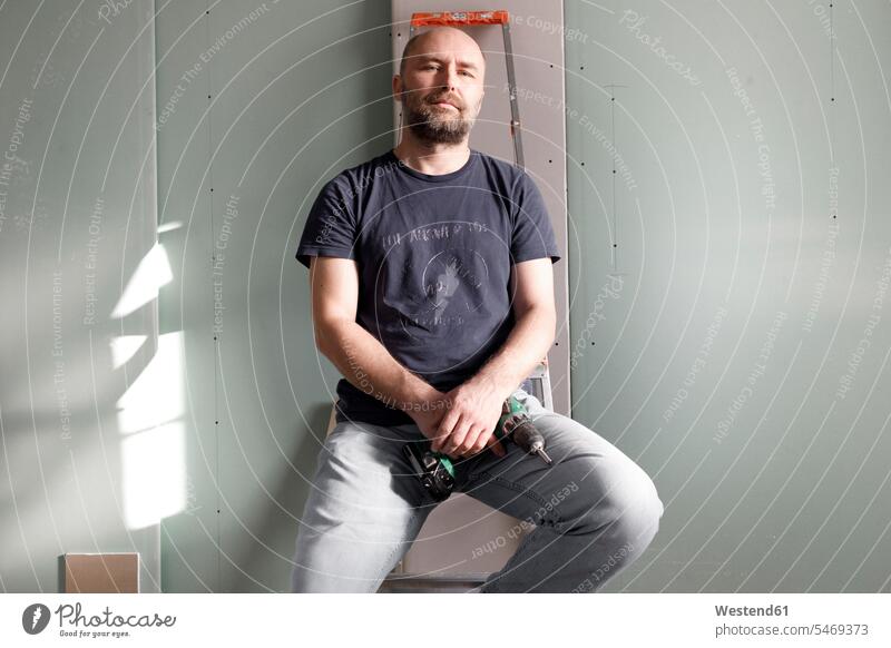 Porträt eines selbstbewussten Mannes, der eine tragbare Bohrmaschine auf einer Baustelle zu Hause hält Leute Menschen People Person Personen Glatzen kahl