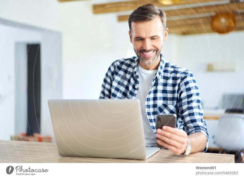 Fröhlicher Mann mit Telefon während der Arbeit am Laptop zu Hause Notebook Laptops Notebooks Zuhause daheim Laptop benutzen Laptop benützen Smartphone iPhone