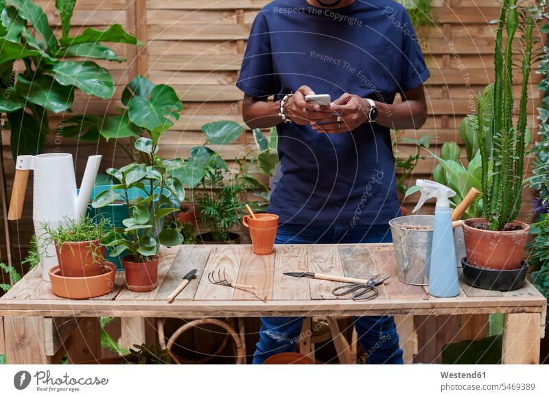 Junger Mann benutzt Smartphone auf seiner Terrasse bei der Gartenarbeit Blumentoepfe Blumentöpfe Tische Holztische Telekommunikation telefonieren Handies Handys