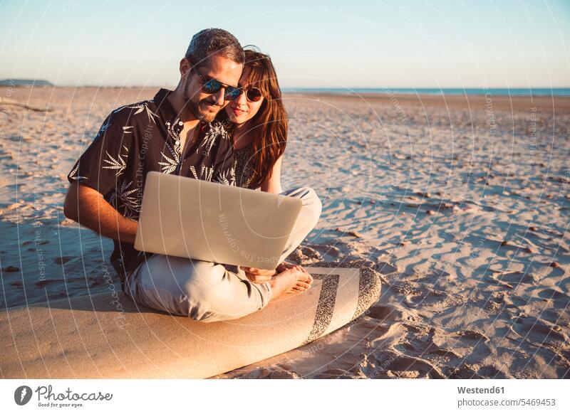 Paar mit Surfbrett sitzt am Strand, mit Laptop Pärchen Paare Partnerschaft Beach Straende Strände Beaches Surfbretter surfboard surfboards Notebook Laptops
