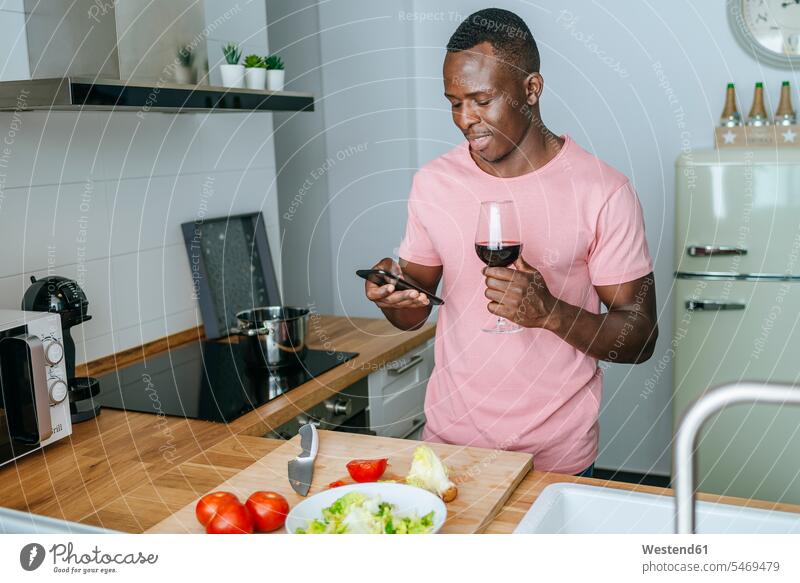 Junger Mann mit Weinglas mit Handy bei der Vorbereitung Salat Leute Menschen People Person Personen Afrikanisch Afrikanische Abstammung dunkelhäutig Farbige