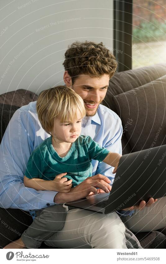 Vater und Sohn schauen sich zu Hause auf der Couch einen Laptop an Notebook Laptops Notebooks sitzen sitzend sitzt Söhne Sofa Couches Liege Sofas ansehen Papas