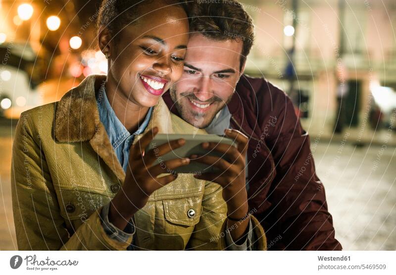 Porträt eines glücklichen jungen Paares, das bei Nacht gemeinsam auf ein Smartphone schaut, Lissabon, Portugal Telekommunikation Handies Handys Mobiltelefon
