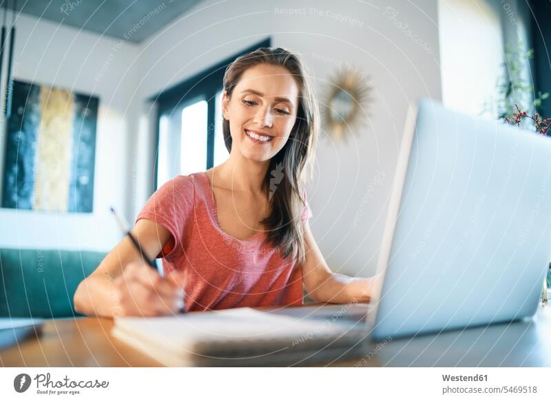 Lächelnde Freiberuflerin schreibt im Notizblock auf dem Schreibtisch im Home-Office Farbaufnahme Farbe Farbfoto Farbphoto Freizeitkleidung Freizeitbekleidung
