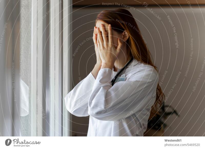 Emotional gestresste junge Ärztin mit Kopf in Händen am Fenster stehend Farbaufnahme Farbe Farbfoto Farbphoto Ärztinnen Doktorin Doktorinnen Arzt Ärzte Doktoren