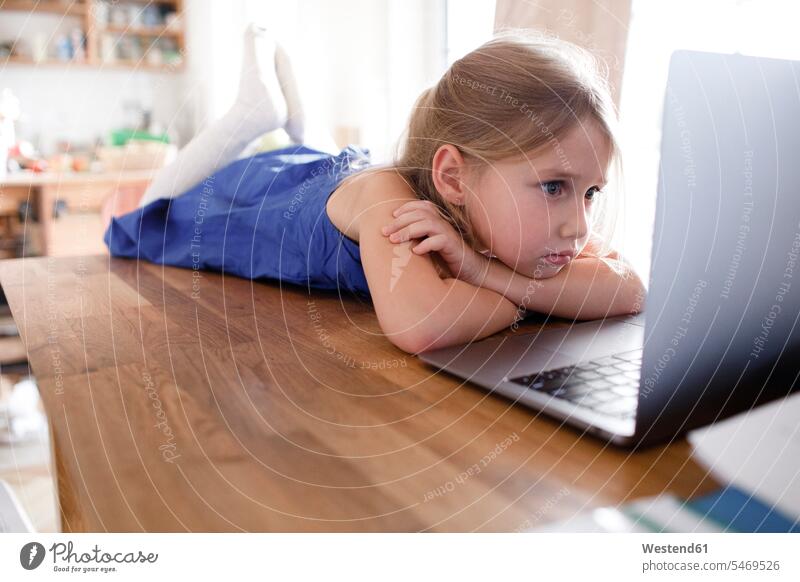 Portrait eines traurigen kleinen Mädchens, das zu Hause auf dem Küchentisch liegt und auf seinen Laptop schaut gefühlvoll Emotionen Empfindung Empfindungen