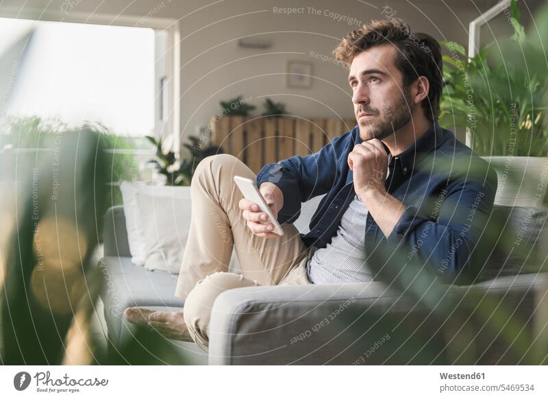 Junger Mann sitzt zu Hause auf der Couch und benutzt sein Smartphone Sofa Couches Liege Sofas Betrachtung Nachsinnen betrachten Kontemplation Unabhängigkeit