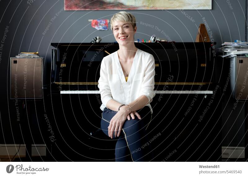 Porträt einer lachenden Frau, die in ihrem Musikzimmer vor einem Klavier sitzt Deutschland Knie anziehen Knie umfassen Freiberufler freiberuflich freie Berufe