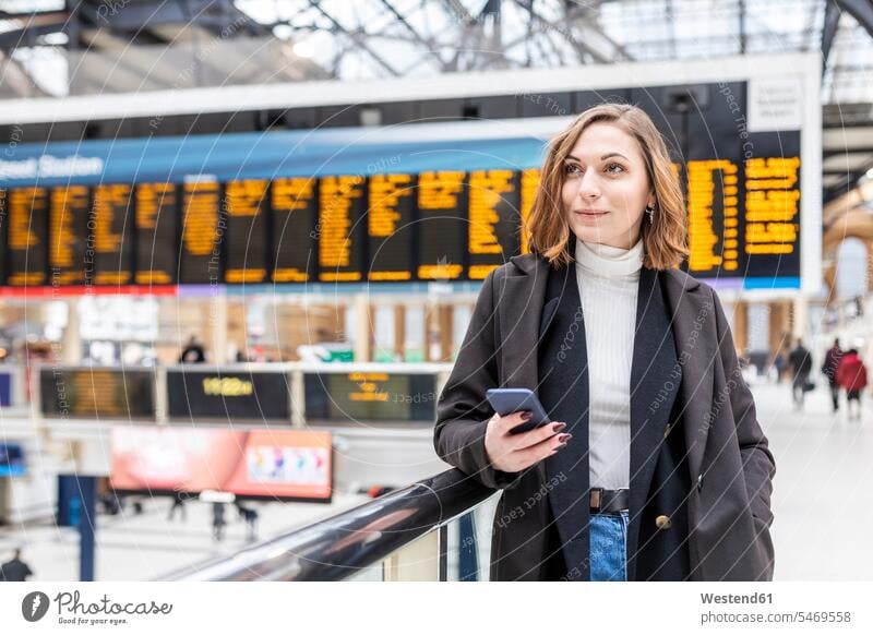 Frau am Bahnhof mit einem Mobiltelefon in der Hand, London, UK Jacken Telekommunikation telefonieren Handies Handys Mobiltelefone zufrieden stehend steht