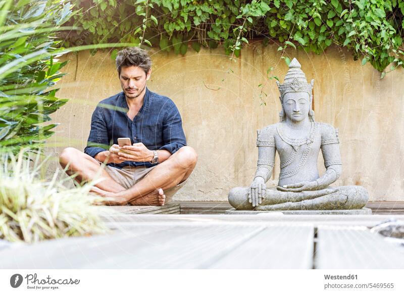 Mann sitzt im Schneidersitz neben einer Buddha-Statue in einem Zen-Garten und benutzt ein Smartphone Handy Mobiltelefon Handies Handys Mobiltelefone SMS