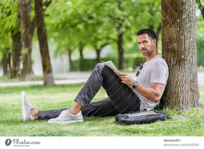 Mann lehnt gegen einen Baum im Park und liest Zeitung Deutschland Seitenblick Blick zur Seite Stadtpark Nachricht Nachrichten Streifen gestreift geringelt Pause