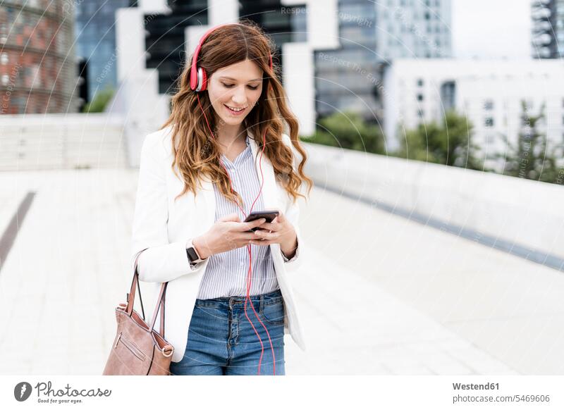 Junge Geschäftsfrau beim Pendeln in der Stadt, mit Smartphone und Kopfhörern unterwegs auf Achse in Bewegung Urban städtisch Urbanität Urbanitaet lächeln
