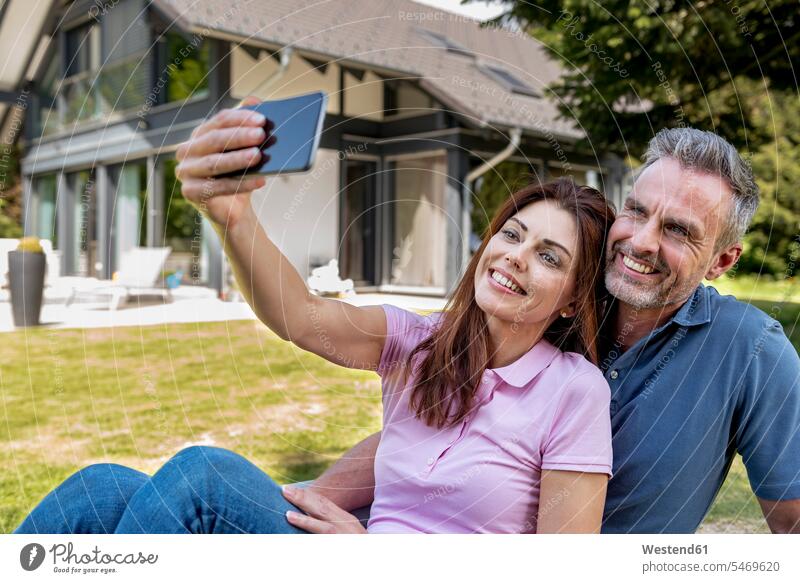 Glückliches Paar sitzt im Garten seines Hauses und macht ein Selfie glücklich glücklich sein glücklichsein Häuser Haeuser Pärchen Paare Partnerschaft Gärten