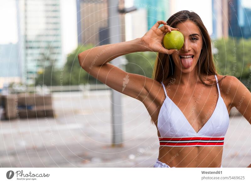 Porträt einer attraktiven, sportlichen jungen Frau, die einen Apfel mit herausgestreckter Zunge hält weiblich Frauen halten Äpfel Aepfel Sportkleidung