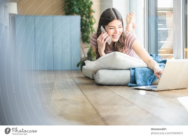 Junge Frau liegt am Fenster zu Hause mit Handy und Laptop Notebook Laptops Notebooks Zuhause daheim lächeln liegen liegend Mobiltelefon Handies Handys