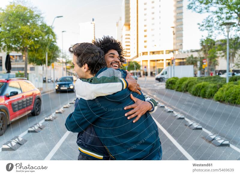 Zwei glückliche Freunde umarmen sich in der Stadt, Barcelona, Spanien Kameradschaft Jacken begruessen Begrüßung grüßen Arm umlegen Umarmung Umarmungen freuen