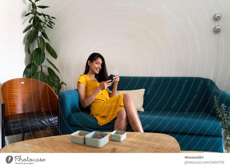 Lächelnde junge Frau sitzt auf Couch und benutzt Mobiltelefon Leute Menschen People Person Personen indisch indische indischer indisches 1 Ein ein Mensch eine