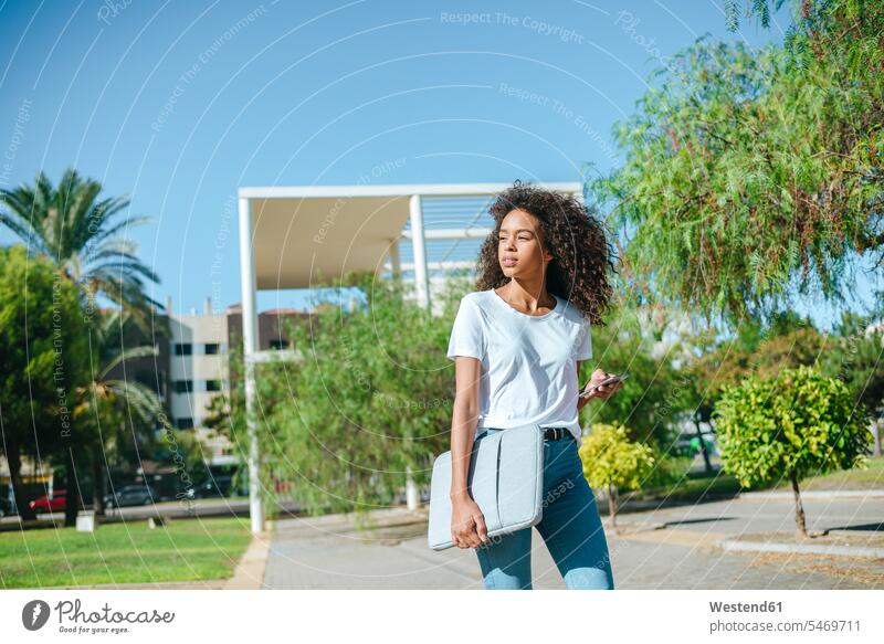 Porträt einer jungen Frau mit Handy und Laptoptasche, die die Straße hinuntergeht und auf ihr Handy schaut Laptop-Tasche weiblich Frauen gehen gehend schauen
