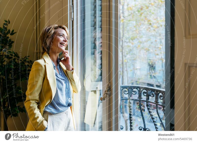 Lächelnde Geschäftsfrau spricht am Smartphone, während sie im Büro durch ein Fenster schaut Farbaufnahme Farbe Farbfoto Farbphoto Innenaufnahme Innenaufnahmen