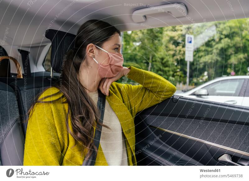 Junge Frau mit Schutzmaske sitzt auf dem Rücksitz eines Autos und schaut aus dem Autofenster Jacken KFZ Verkehrsmittel Automobil PKW PKWs Wagen Farben Farbtoene