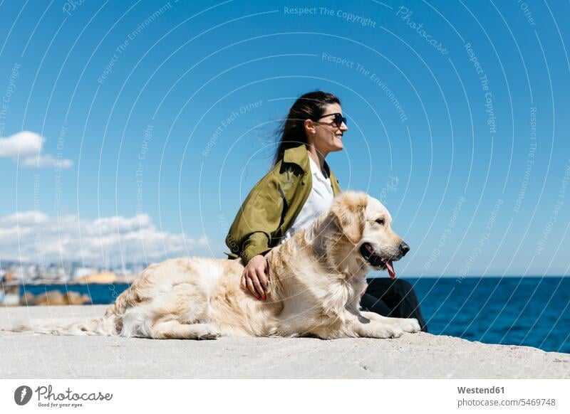 Glückliche Frau sitzt neben ihrem Labrador Retriever am Kai und schaut auf das Meer Hundebesitzer Horizont Horizonte Textfreiraum gehen gehend geht Gassi gehen