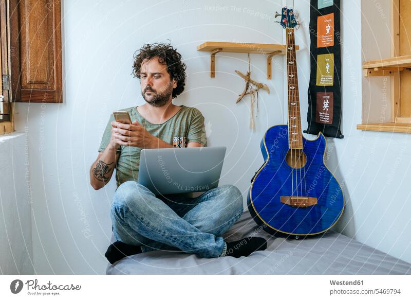 Mann in seinem Zimmer mit Mobiltelefon, Laptop und Bassgitarre Smartphone iPhone Smartphones Männer männlich Freizeit Muße Musik Notebook Laptops Notebooks