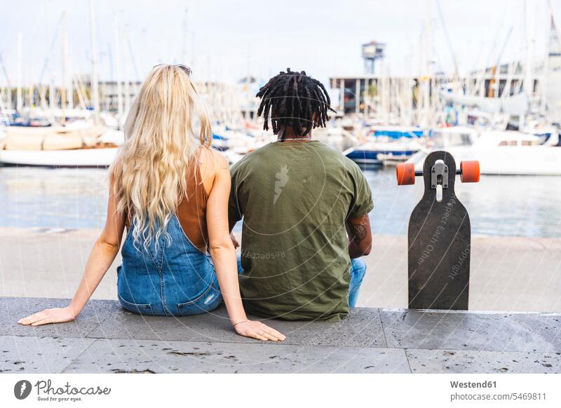 Spanien, Barcelona, Rückenansicht eines multikulturellen jungen Paares nebeneinander sitzend mit Longboard im Yachthafen Jachthafen Jachthäfen Yachthaefen