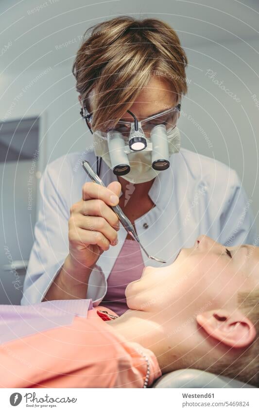Zahnarzt, der die Zähne eines Jungen untersucht, mit Kopflupe und Dentalinstrument Patient Kranke Kranker Patienten Zahnarztinstrumente